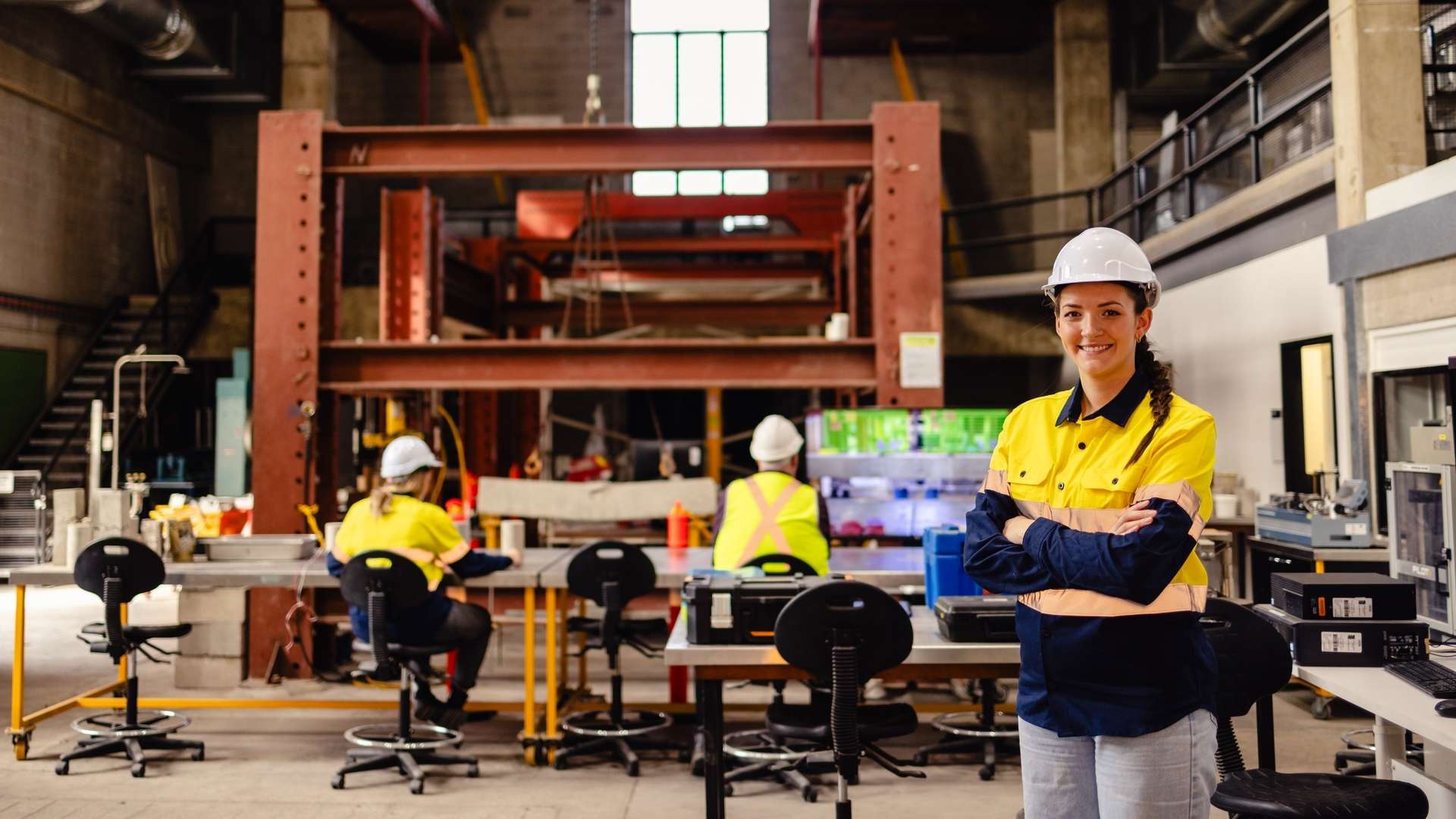 A CQU engineering student wearing high vis and a hard hat stands in front of an engineering workshop.