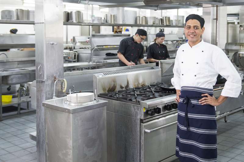 A hospitality student in a chefs uniform standing in a large professional kitchen