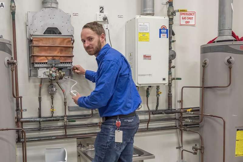 Image of man in blue shirt and jeans standing in front of plumbing training equipment