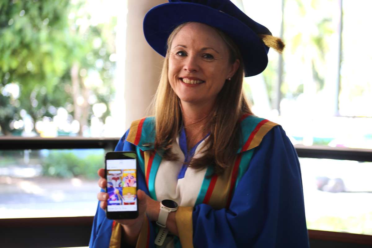 Dr Gitte Galea stands in graduation robes with a phone showing a Queensland Health instagram meme.
