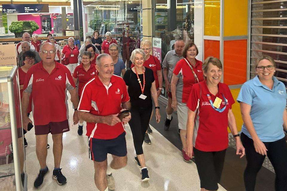 A large group of older men and women, mainly wearing a red uniform, walk as a group through a shopping centre.