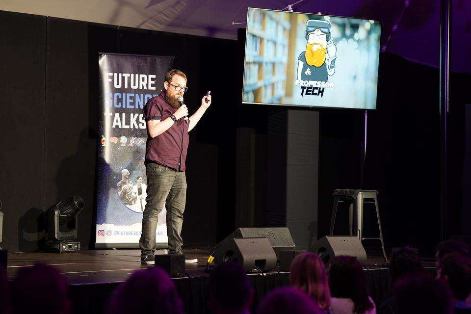 Michael Cowling stands on Brisbane Comedy Festival stage, screens show his Professor Tech avatar.