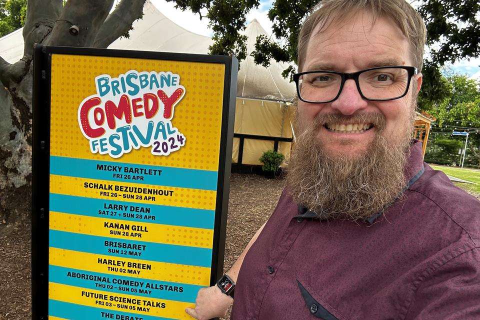 Michael Cowling stands outdoors in front of Brisbane Comedy Festival sign, he points to Future Science Talks listing.