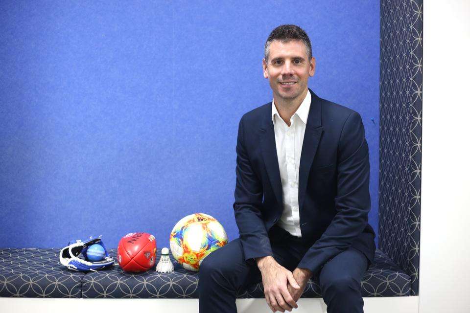 Fabio Serpiello wears a suit, sits indoors on a bench next to sporting equipment.