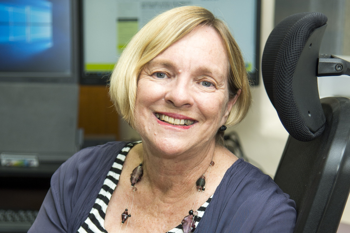 Woman with short blonde bob hair cut sits in front of PC screens and smiles at camera. She is wearing pink lipstick with teeth showing, a long beaded necklace and a light cardigan over a black and white horizontally striped shirt