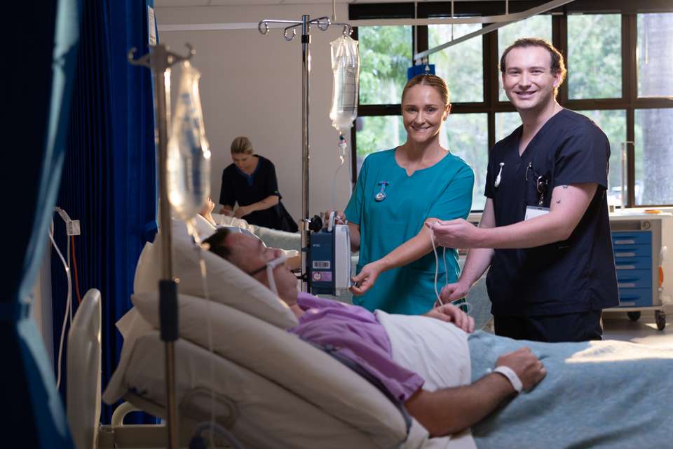 Two nursing students caring for a patient who is in bed in a realistic hospital setting.