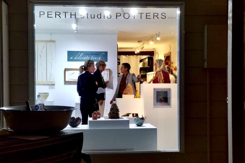 A view from the outside of a lit art gallery window at night; people inside are talking and viewing the art.