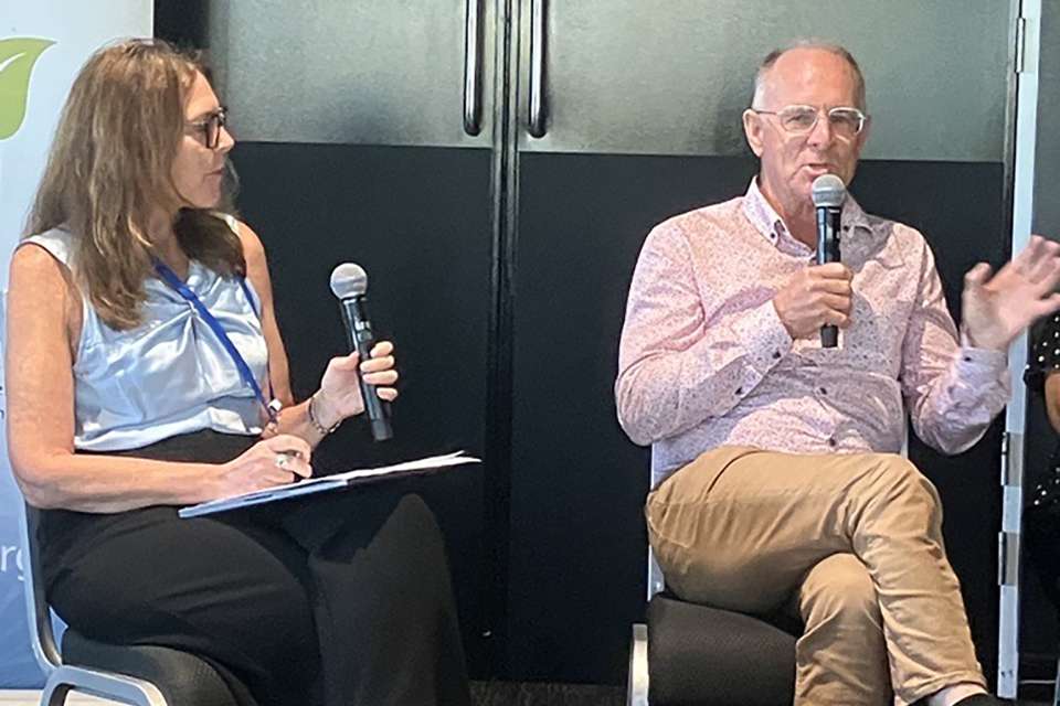 Image of John Rolfe holding a microphone with a woman seated beside him on a microphone