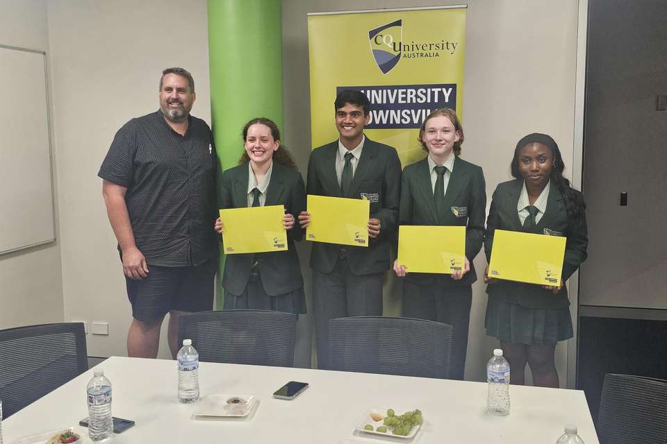 Four students standing with a teacher in front of a green CQU banner