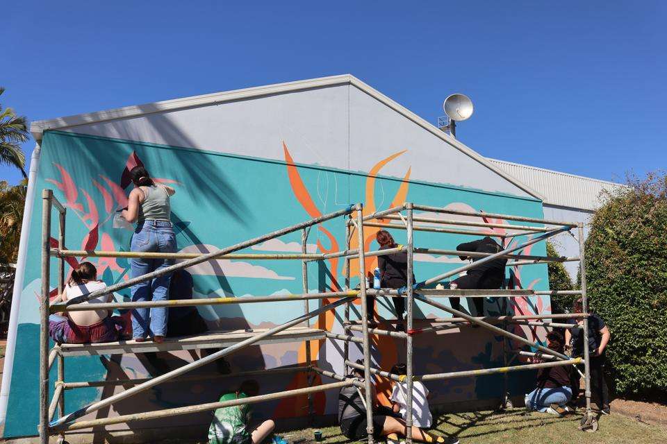 A group of students paint a brightly coloured mural featuring a kangaroo on a building wall at the Bundaberg campus. They sit and stand on scaffolding while doing this.