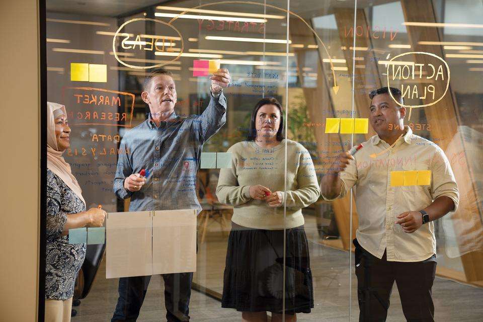 Four people stand behind a glass window in an office space, and strategise by writing plans and posting post-it notes on the glass.