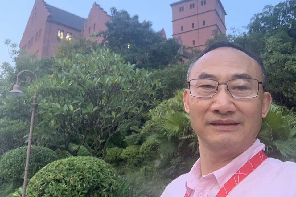 Hong Shen stands outdoors, in front of a towering building and trees.