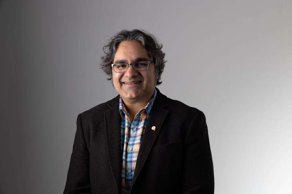 Professor Anjum Naweed stands indoors smiling, wears a dark jacket with a lightbulb pin on the lapel.