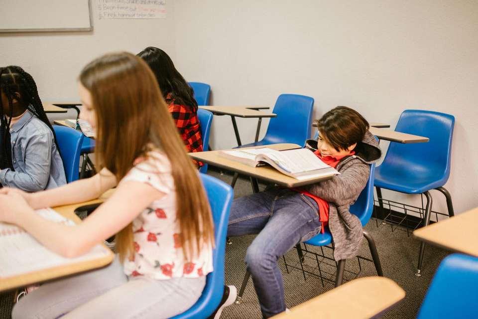 Students sit in a classroom setting, with the focus on a boy slumped at his desk in front of an open textbook.