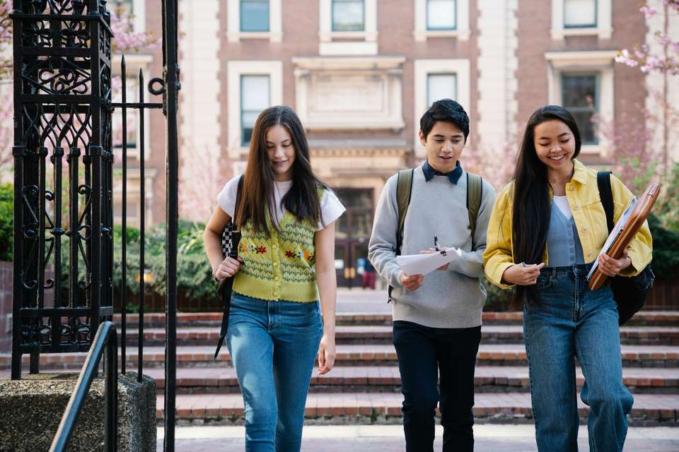 Three students walk through a cast iron gate with school grounds in the background. They are smiling and appear to be in mid-conversation, carrying their books and bags as they walk together.