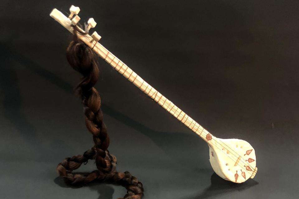 A sculpture of a sitar with a long hair braid attached on a grey background