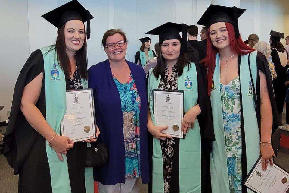 Four smiling women stand indoors, three wearing mortarboards and stoles and holding testamurs.