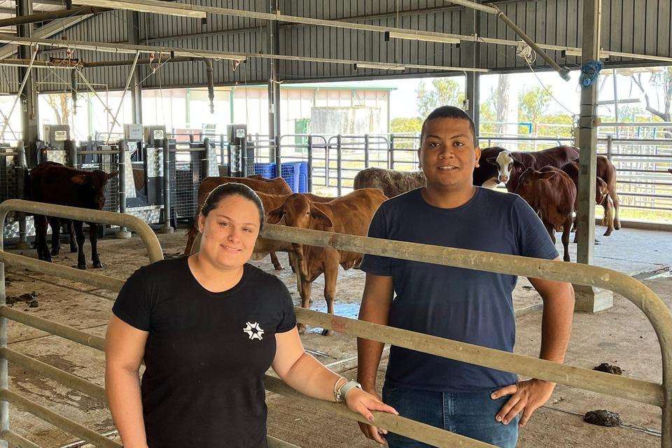 Smiling woman wearing black t-shirt that has a white round-shaped logo on the left breast stands in front of a cattle yard that has cows in the background alongside a man wearing a plain blue t-shirt and jeans with hands on hips also looking and smiling directly at the camera