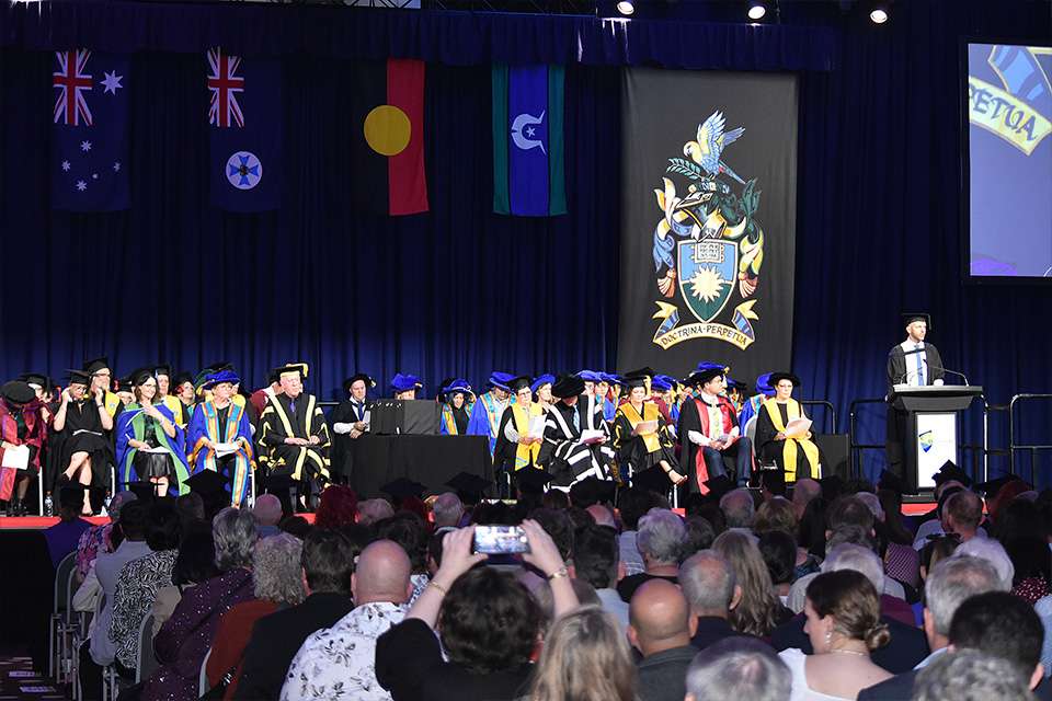 A wide image of people seated on stage in ceremonial graduation garb and attendees in the foreground