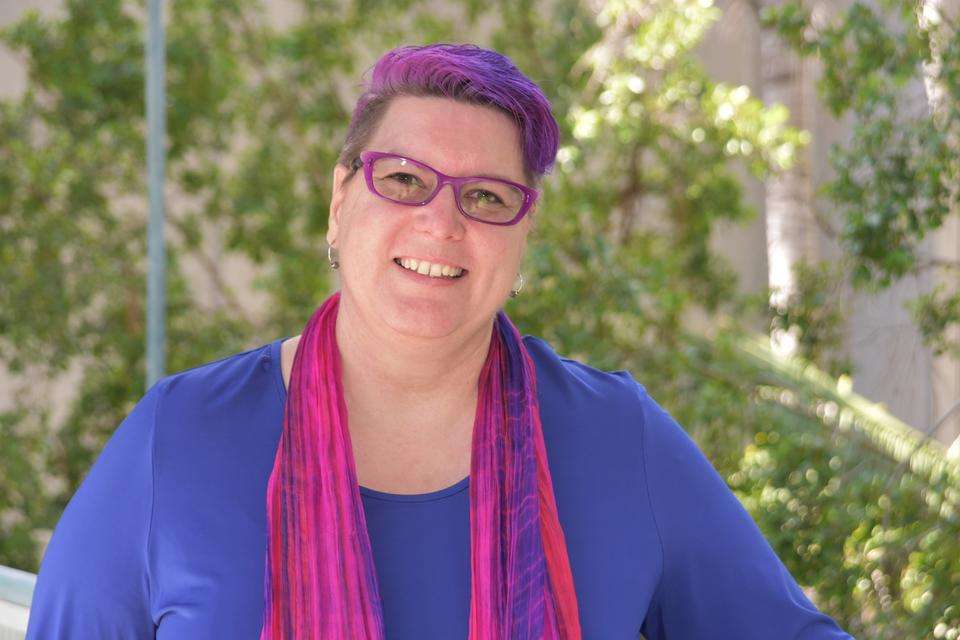 Woman with short pink and purple hair wearing purple reading glasses, a bright blue blouse and pink scarfe