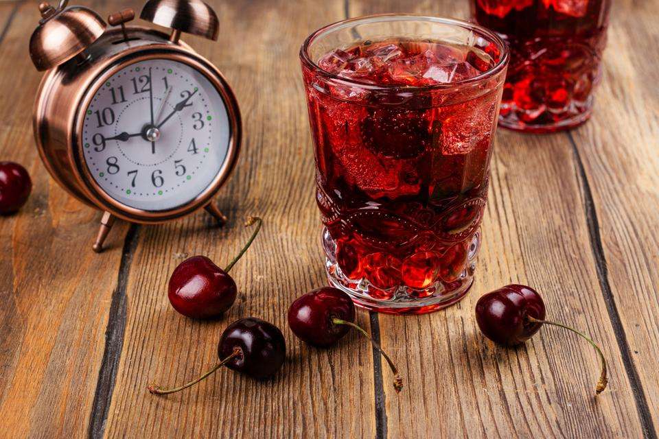 An analogue clock with a red drink in the foreground and cherries on the table