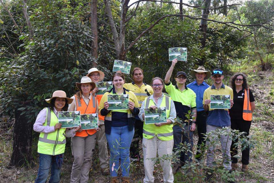 A group of people stand together smiling at the camera and holding certificates of appreciation, standing amongst native bushland. They are wearing protective clothing and some wear high-vis.