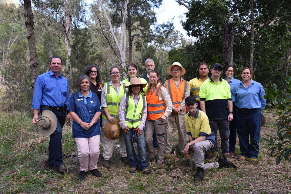 A group of people stand together smiling at the camera, standing amongst native bushland. They are wearing protective clothing and some wear high-vis.