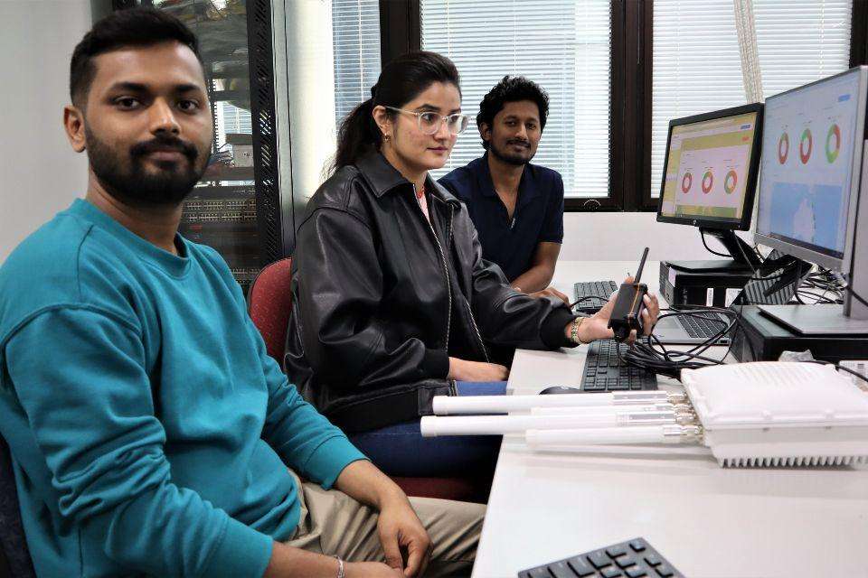 Three students sit at a desk with IoT monitoring technology and a computer dashboard display.