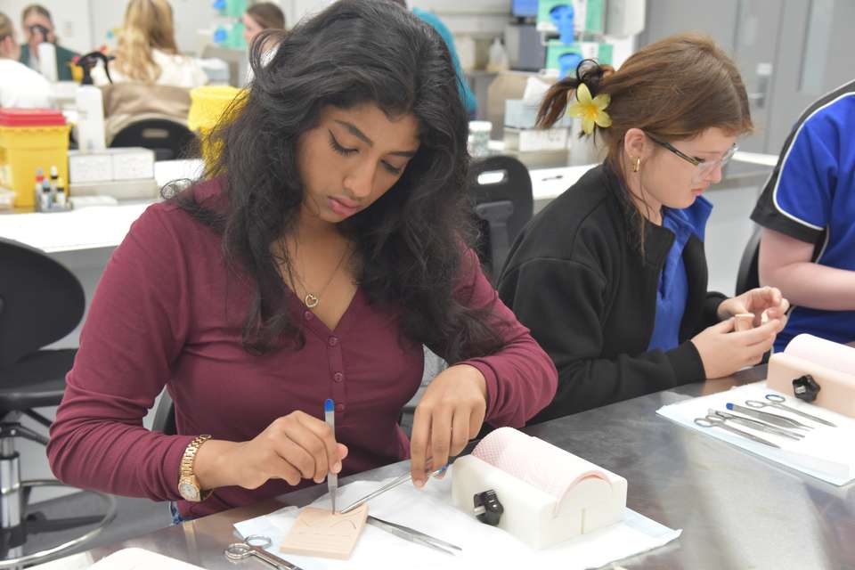 A high school student sits at a lab table with medical suturing tools and practices suturing on dummy skin.