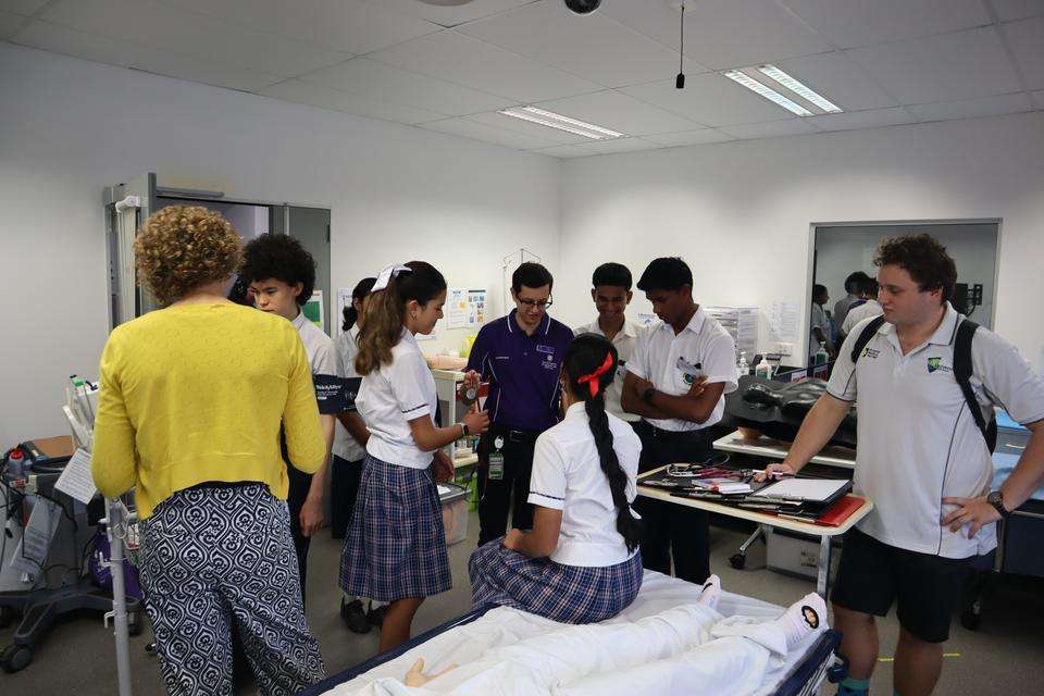 A large group of students in school uniforms practice taking each others blood pressure using a blood pressure cuff and stethoscope. One student sits on a mock hospital bed while instructors guide the others on how to use the equipment and check other vital signs.