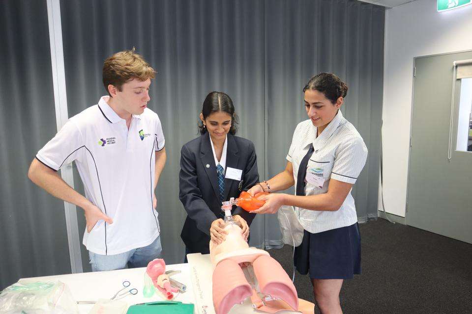 A young man instructs two young women in a medical simulation scenario on airway management. They are practicing on a medical mannequin and are using an oxygen mask and bag.