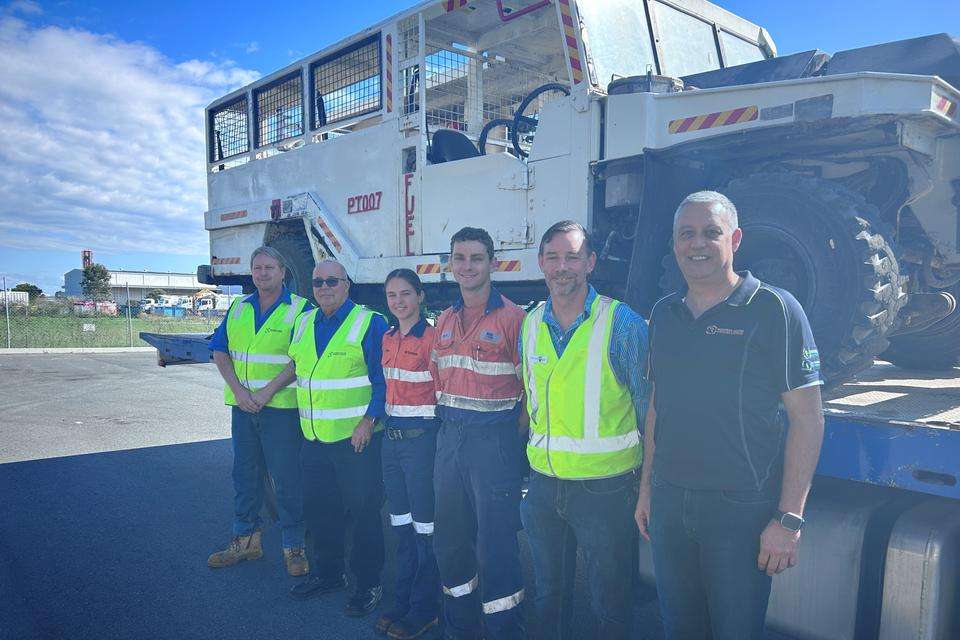 Six people wearing high vis stand smiling at the camera, in front of a vehicle atop a truck tray