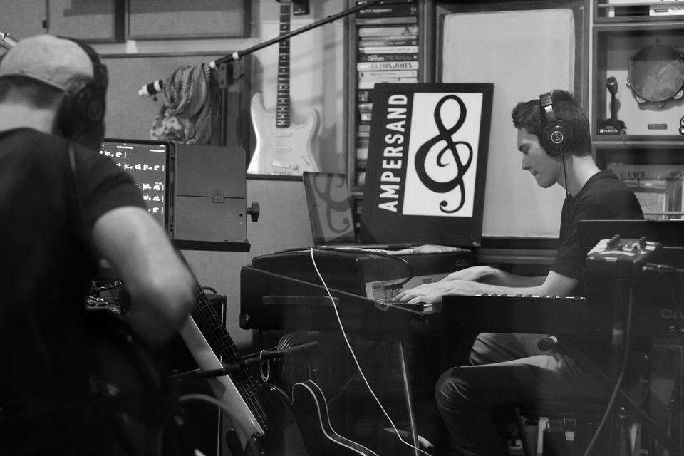 A black and white image of a man playing guitar, facing away from the camera, and a man wearing headphones playing a keyboard. A sign in the studio reads: Ampersand.