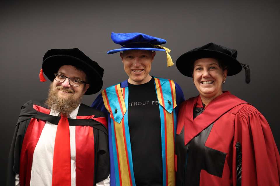 Three people stand dressed in colourful graduation regalia, smiling at the camera