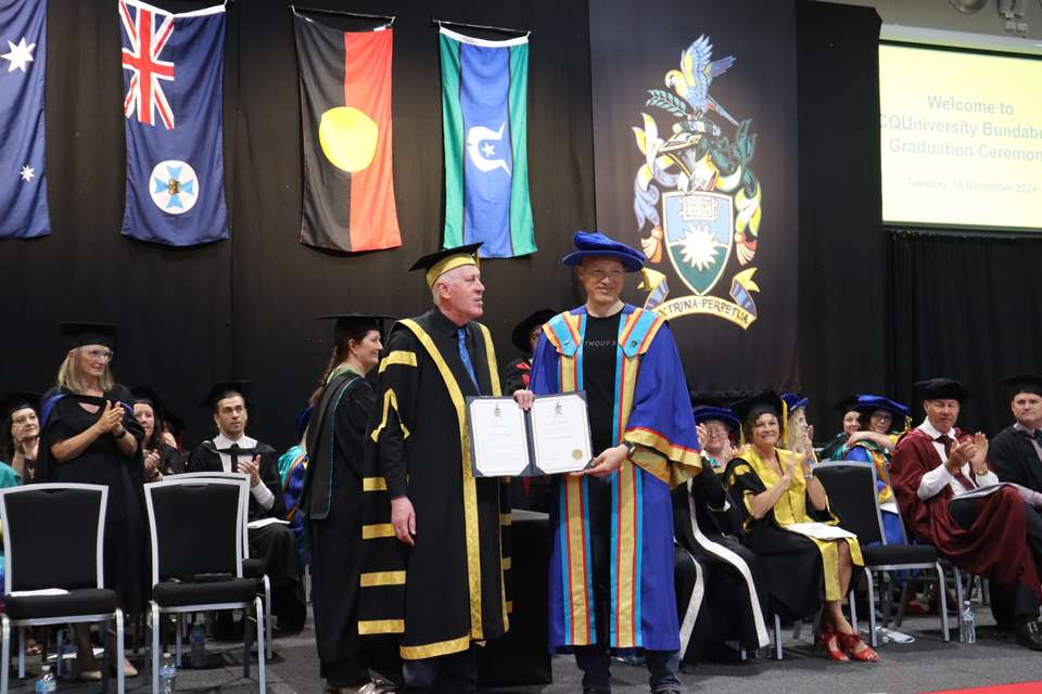 A PhD graduate stands on stage with the CQU Chancellor to receive his testamur. Academics applaud onstage in the background against a backdrop of flags and the University crest.