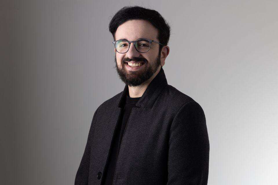 A smiling man with dark hair and a beard, wearing glasses and a black tshirt and jacket.