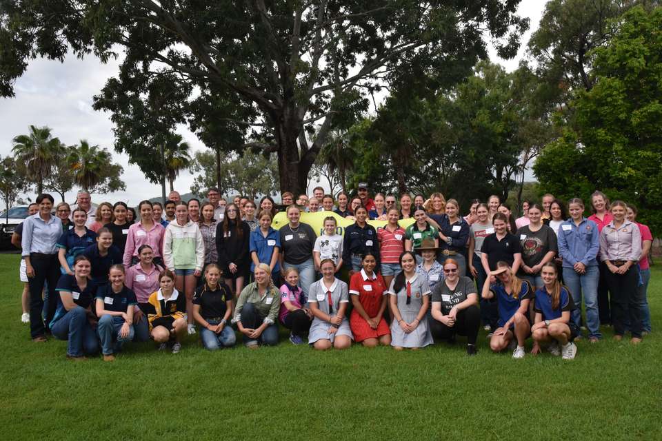 A group of 50 high school students and their teachers, mentors and CQU academics stand under a large tree outside, gathered around a CQU-branded bull statue.