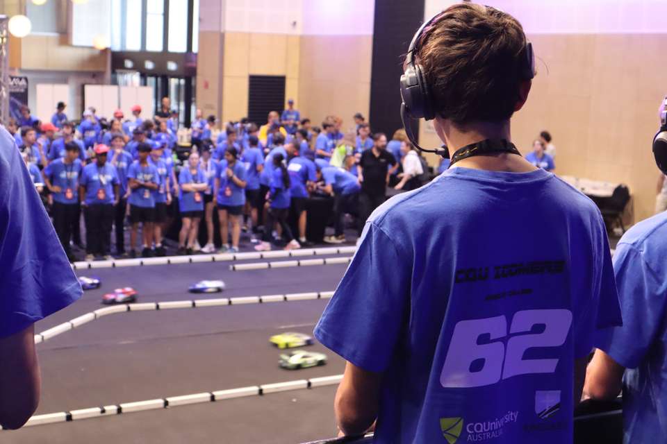 A CQU-sponsored Hydrogen Grand Prix competitor stands onstage in the drivers' pit, overlooking the race track as cars zoom by and teammates look on in the background