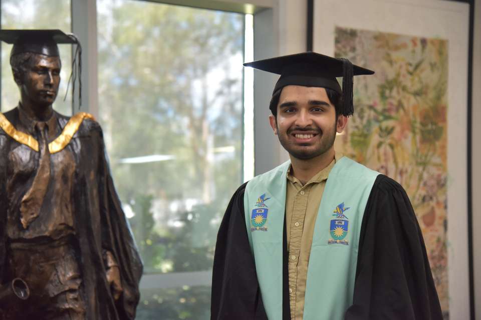 A young graduate stands smiling in graduation regalia, a floral painting and graduate statue feature in the background.
