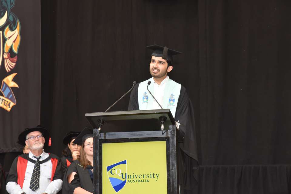 A young graduate in regalia stands on stage at a podium delivering a speech. A backdrop of black and part of the CQU crest, along with seated academics feature in the background.