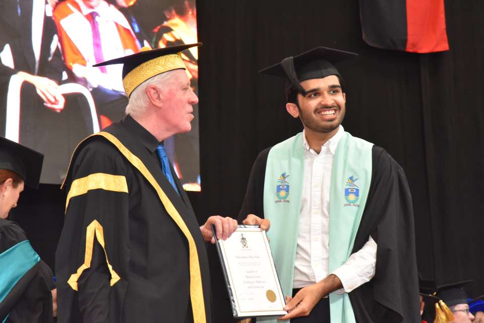 A young graduate smiles as he stands with the CQU Chancellor to receive his testamur for the Bachelor of Medical Science (Pathway to Medicine)