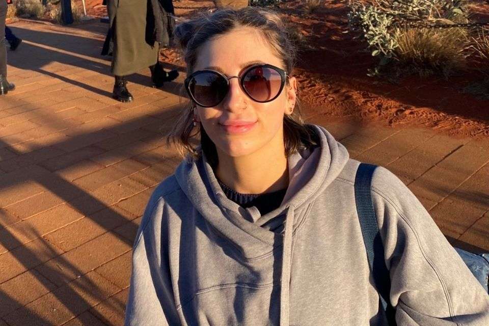 A young woman wearing a grey jumper adn sunglasses smiles in sunny outdoor location.