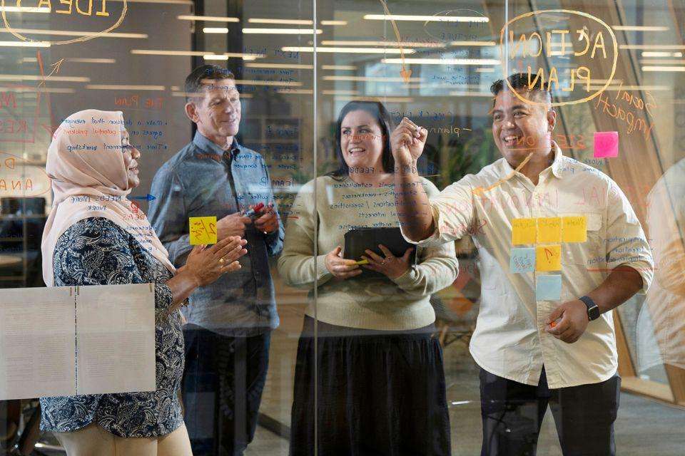 Two women and two men stand behind a window, writing a plan and adding post-it notes to to the glass.