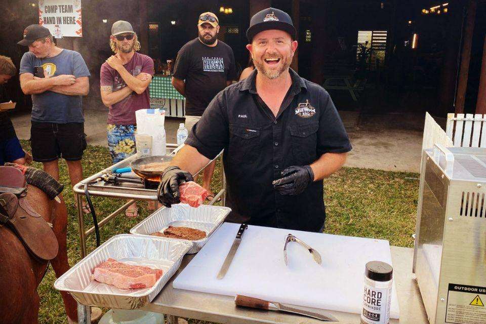 A man wearing a cap holds a piece of beef at an outdoors BBQ prep table.