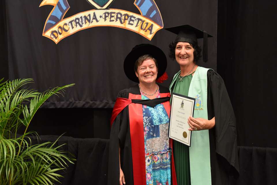 Dr Jay Deagon and Evette Lonergan stand in graduation robes on a stage, smiling.