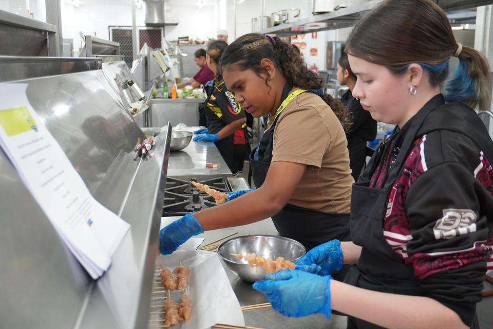 Two high school girls work together in a commercial kitchen making chicken skewers