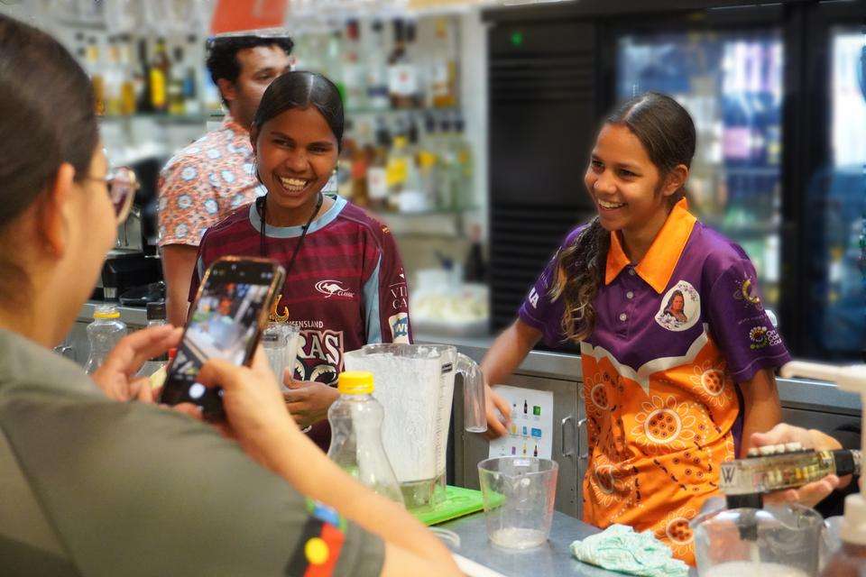 Two high schools stand behind a restaurant training bar, smiling at the camera, with an array of drink-making equipment in front of them on the bench. Someone stands in front of them taking a photo