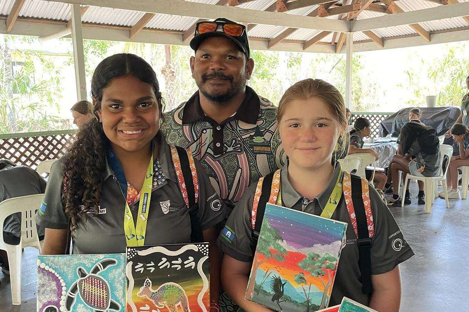 Two Indigenous Girls Academy Leadership team members girls holding up their paintings with a male mentor in the background