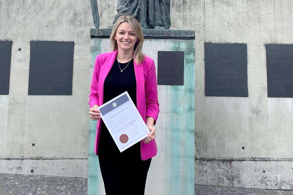 Blonde woman wearing black dress and pick jacket holding a testamur and standing in front of a statue