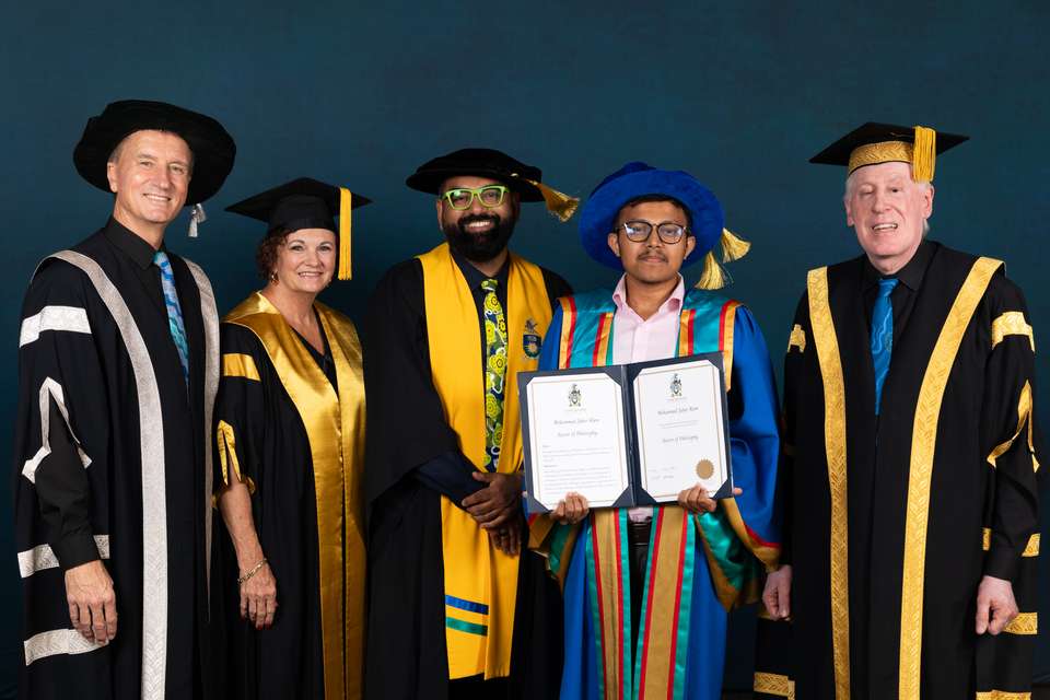 Five people stand smiling for the photo, dressed in colourful academic regalia. People include university leadership, an academic, and a PhD graduate who holds his testamur proudly.